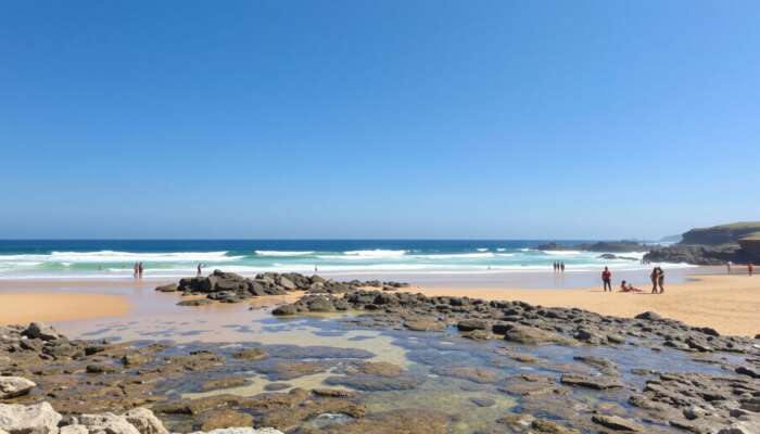 A sunny beach with clear waves, golden sands perfect for sunbathing, rocky outcrops for exploration, accessible ramps, and diverse groups enjoying the day under a blue sky.